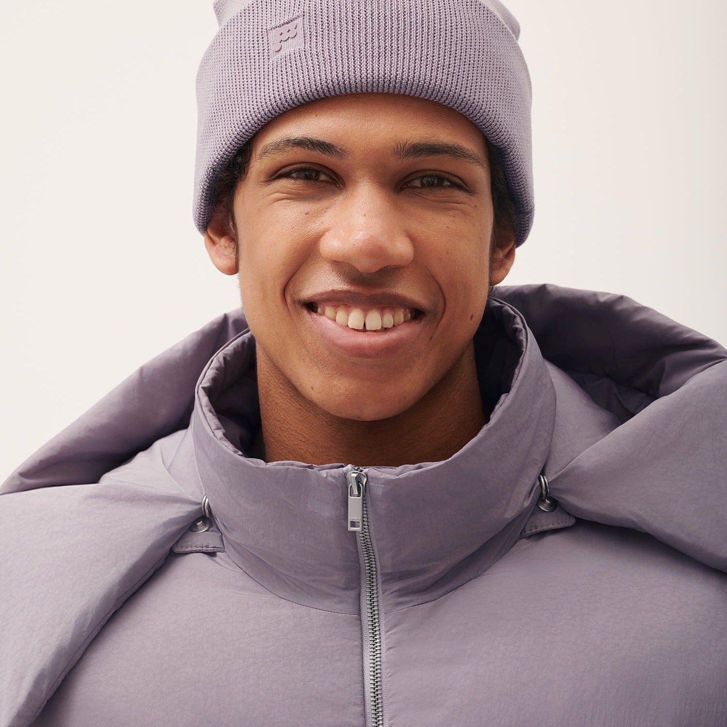 Smiling man in Pangaia lilac ribbed knit beanie with P logo, matching quilted puffer jacket, high zippered collar