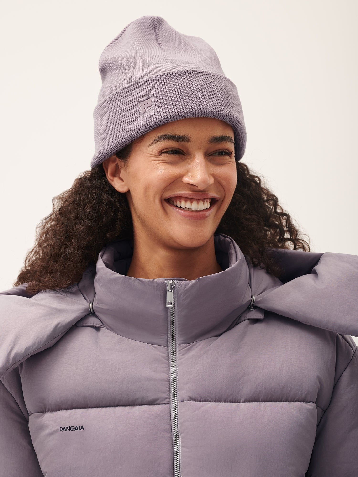 Smiling model in Pangaia lavender ribbed knit beanie and purple puffer jacket high collar full zip winter style