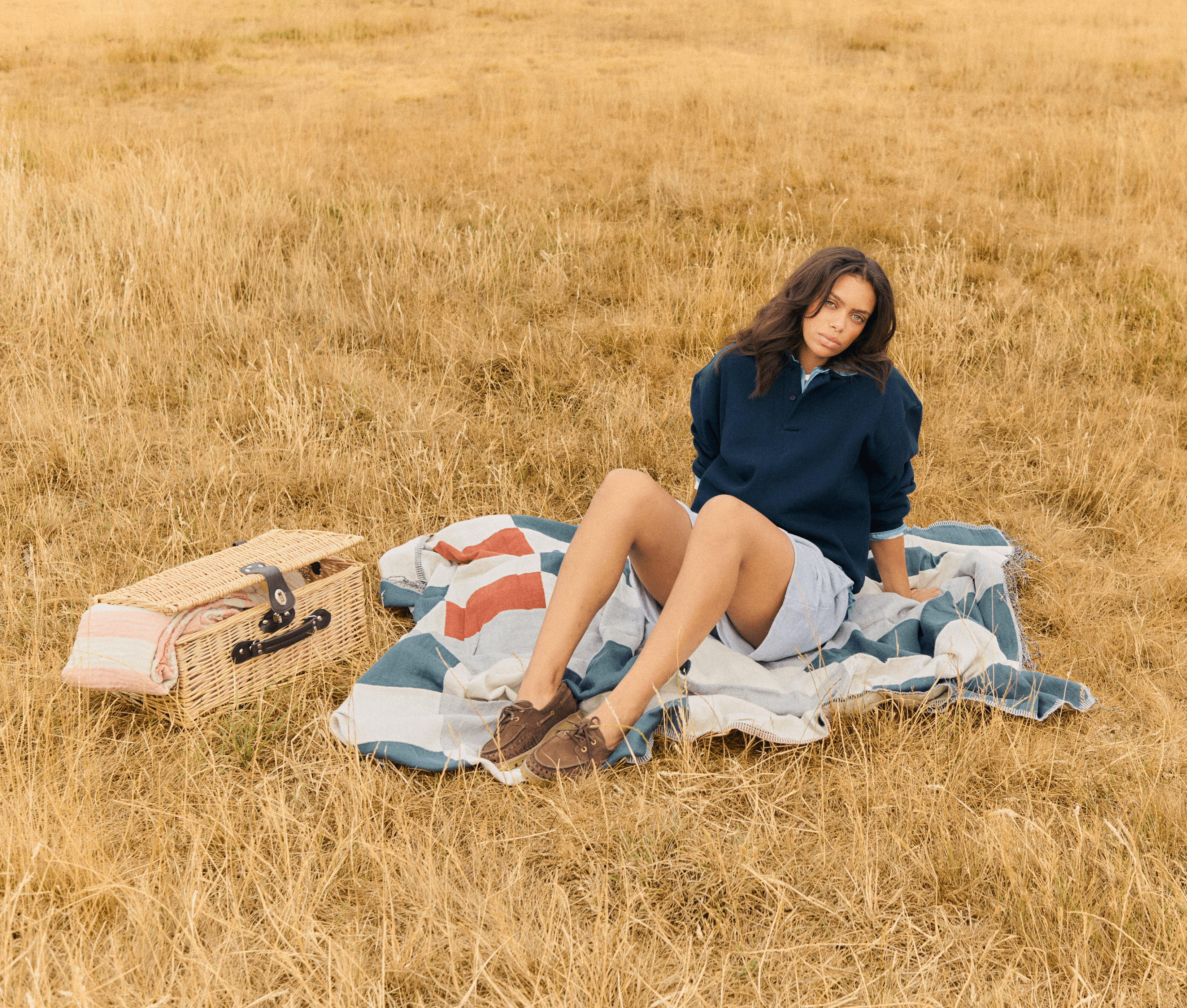Model in navy long-sleeve collared rugby shirt, light blue striped linen shorts, brown leather boat shoes, picnic blanket