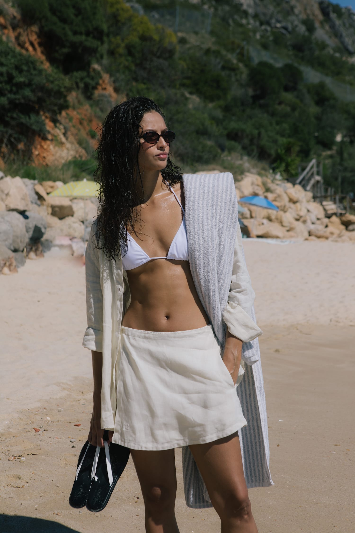 Woman wearing white triangle bikini top, cream linen blend mini skirt, open cream button-up shirt, light blue striped towel