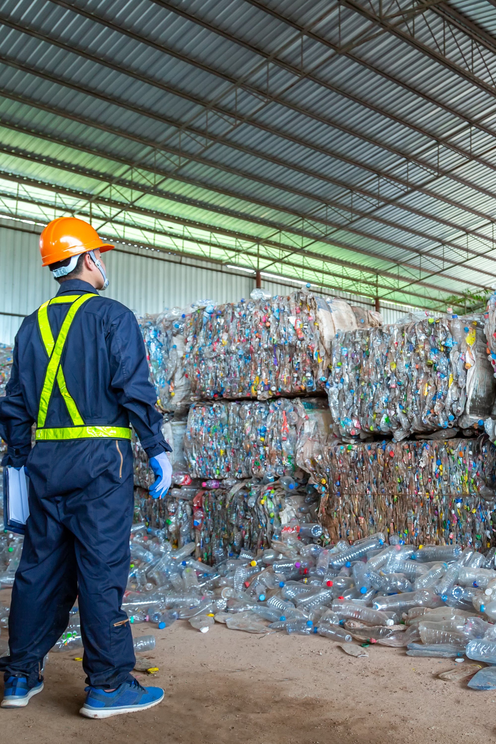 Dark blue work coveralls with high-visibility yellow reflective strips and waist band, orange hard hat, blue gloves, back view in recycling plant