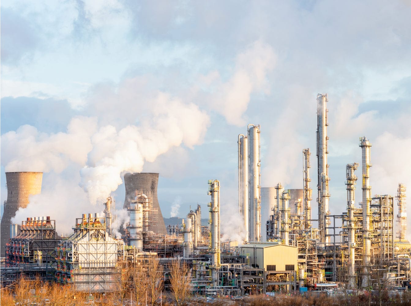 Industrial petrochemical plant with grey and orange cooling towers, dense network of pipes, tanks, and smokestacks releasing steam